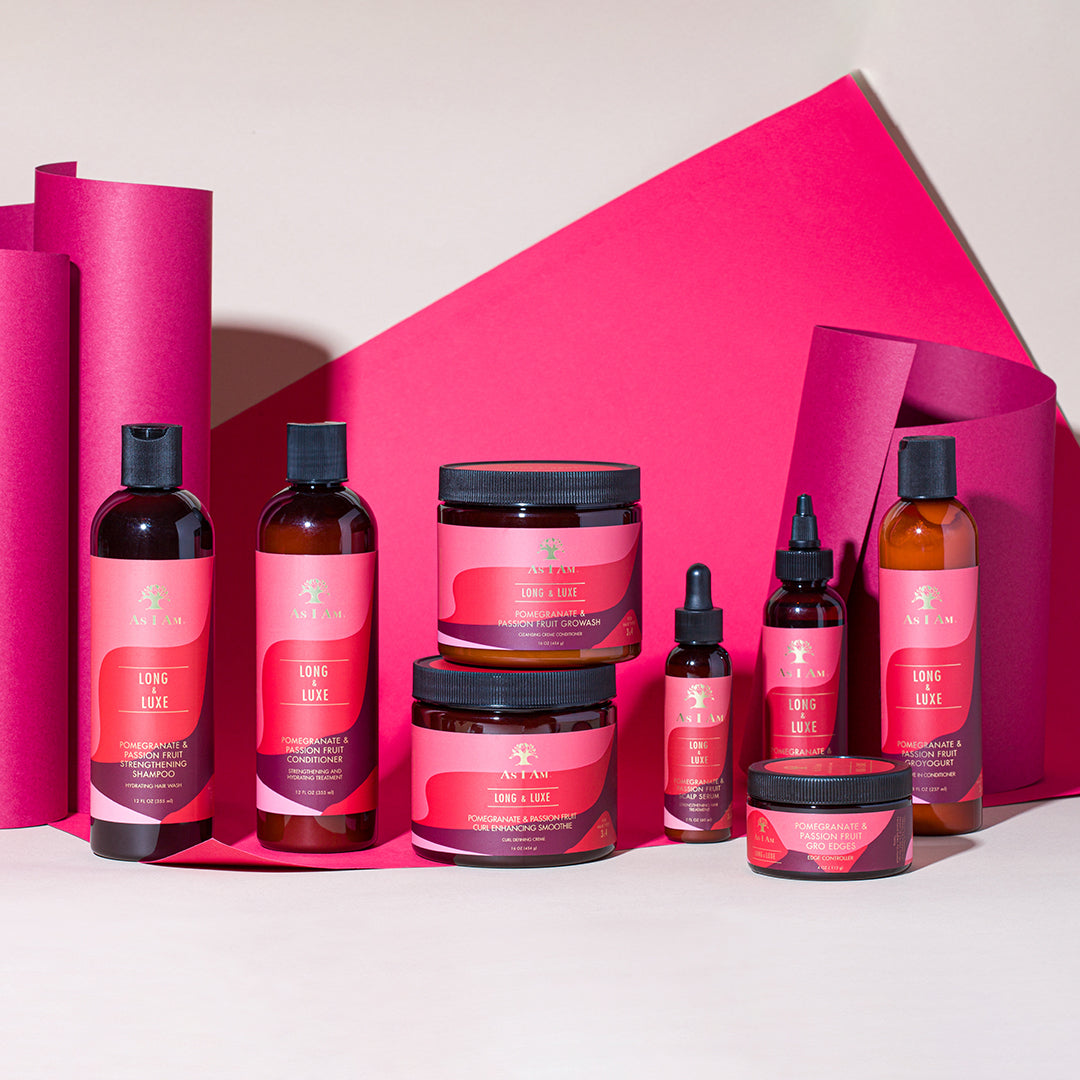 En samling af produkter fra As I Am Long & Luxe-serien, herunder shampoo, balsam, leave-in conditioner, olier og krukker, arrangeret foran en lyserød baggrund.