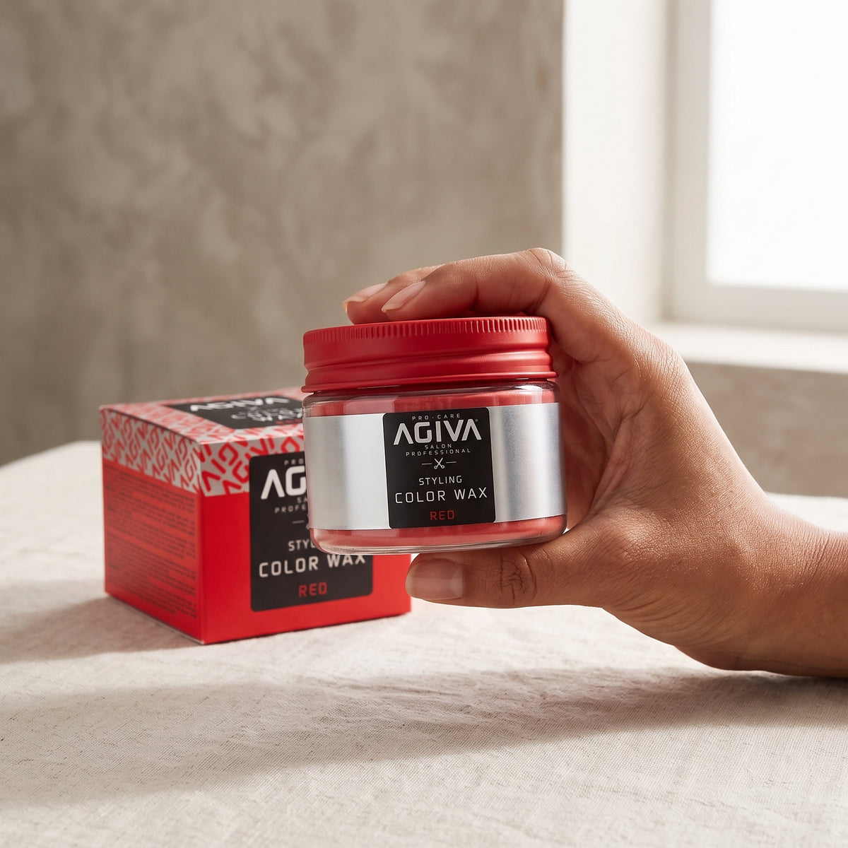 Hånd holder AGIVA Styling Color Wax Red, rød hårvoks i glas med rødt låg, ved siden af den røde emballage.