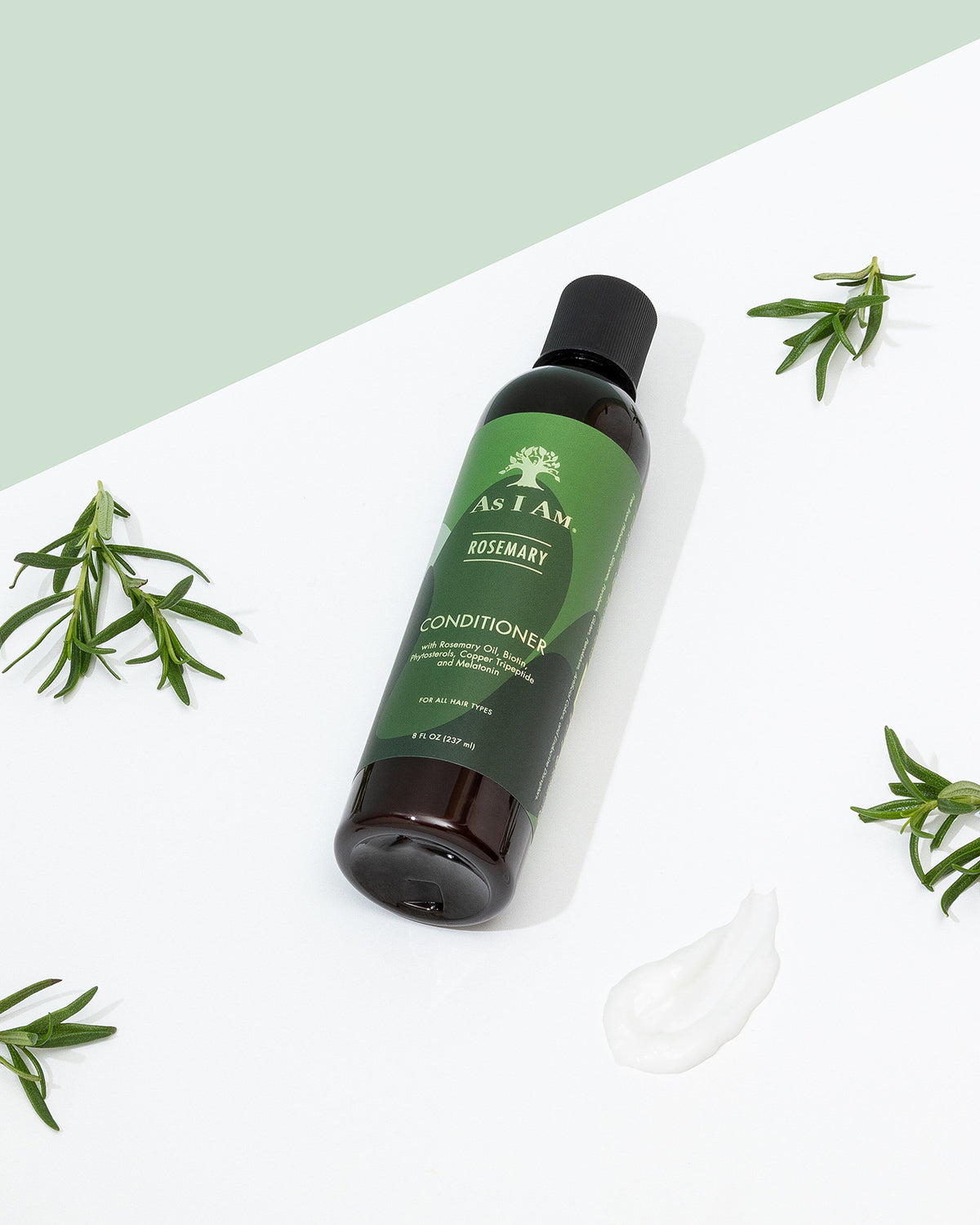 En flaske As I Am Rosemary Conditioner (237 ml) ligger diagonalt på en hvid overflade med rosmarinkviste spredt omkring og en klat balsam.