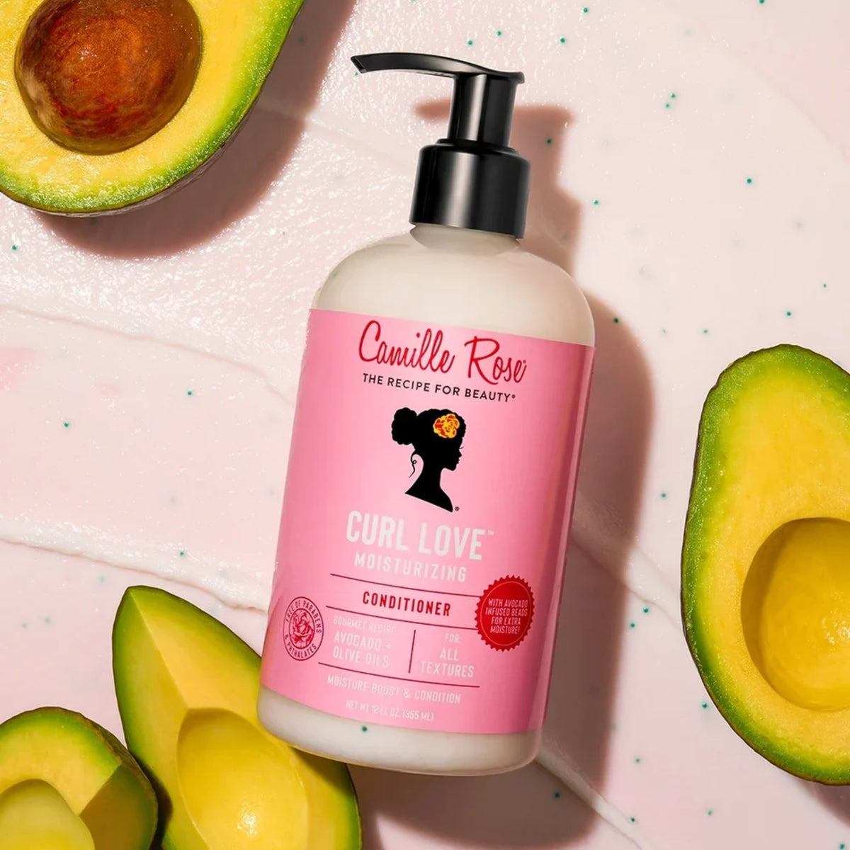 Camille Rose Curl Love Moisturizing Conditioner i pumpeflaske omgivet af skårne avocadoer på lyserød baggrund.
