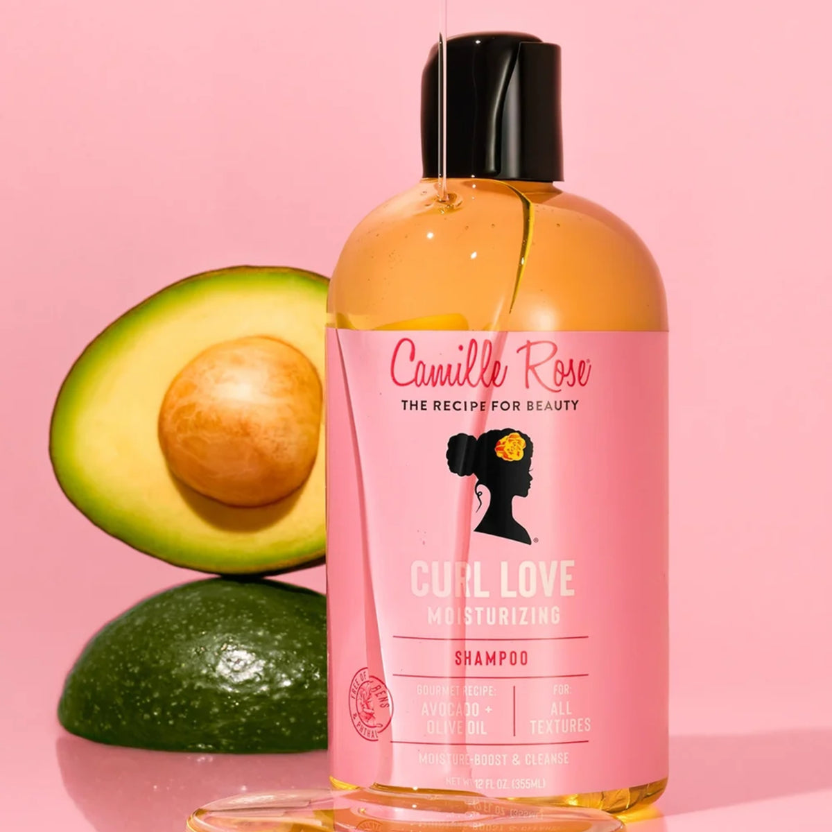 Camille Rose Curl Love Moisturizing Shampoo-flaske med avocado og olivenolie foran skåret avokado på lyserød baggrund.