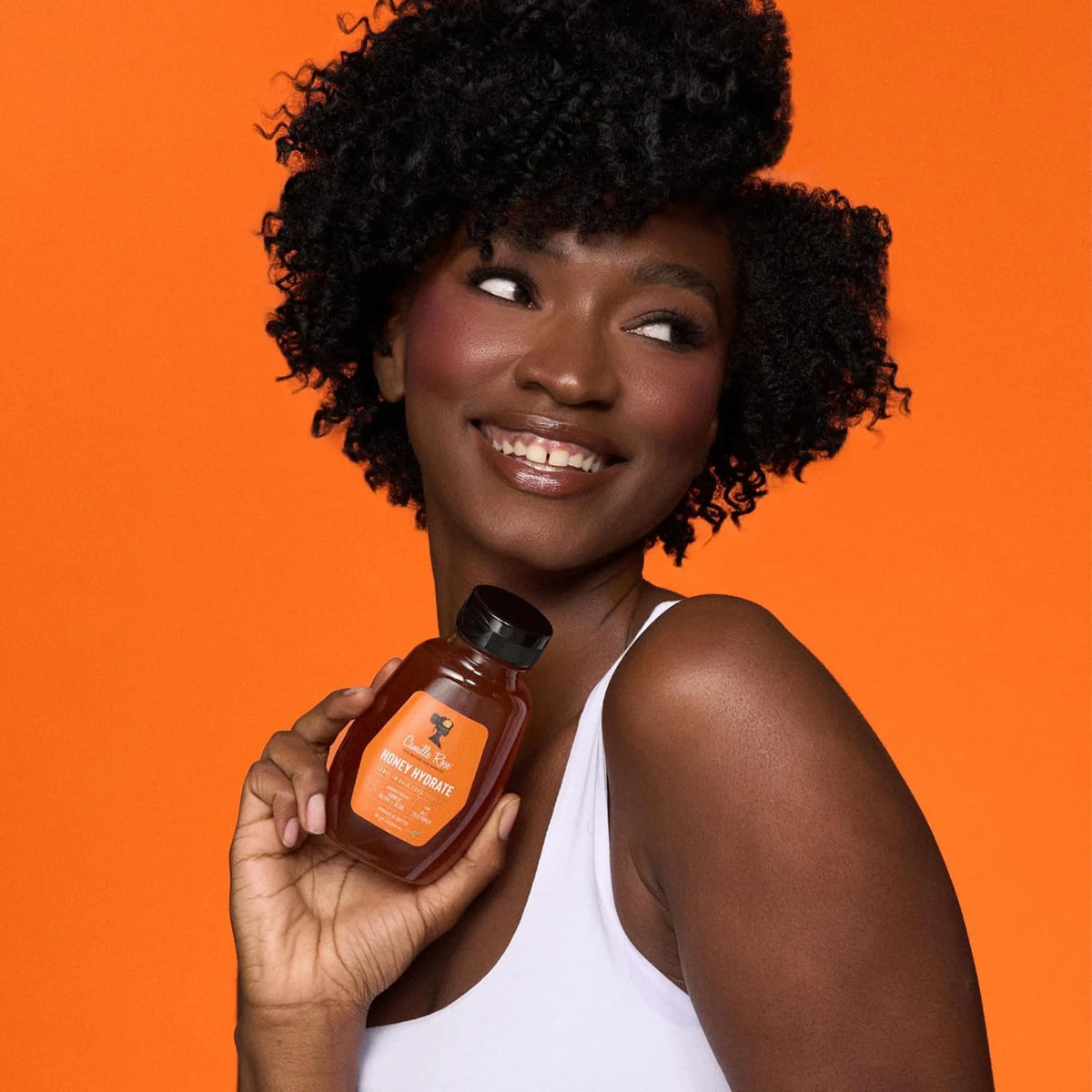 Smilende kvinde med naturligt krøllet hår holder Camille Rose Honey Hydrate hårbalsam foran orange baggrund.