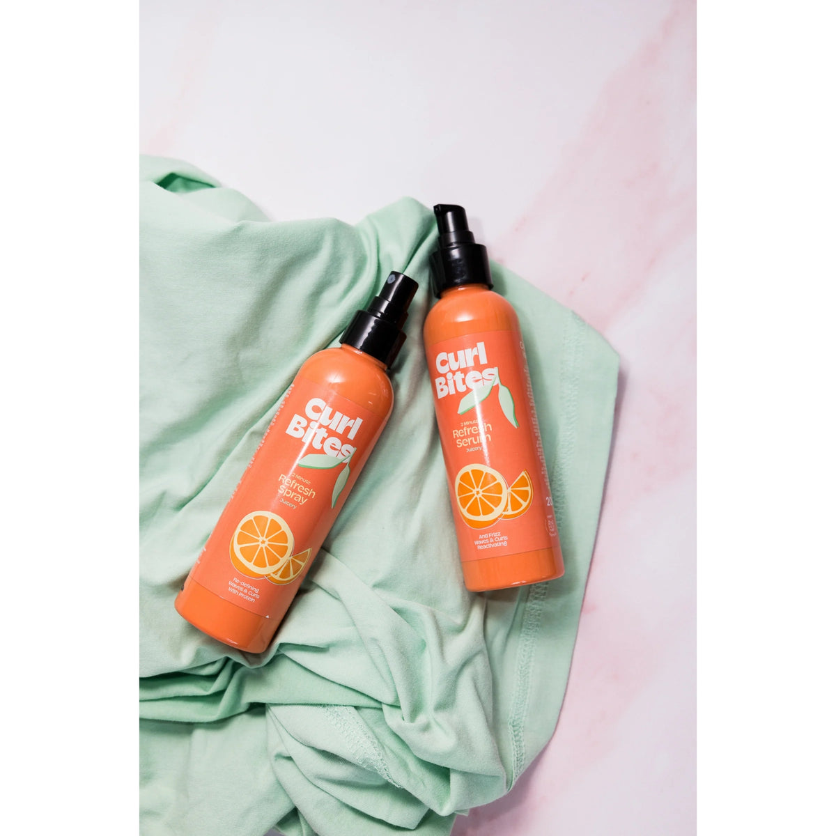 Curl Bites Refresh Spray og Refresh Serum i orange flasker med sort pumpelåg, placeret på mintgrøn stofbaggrund.