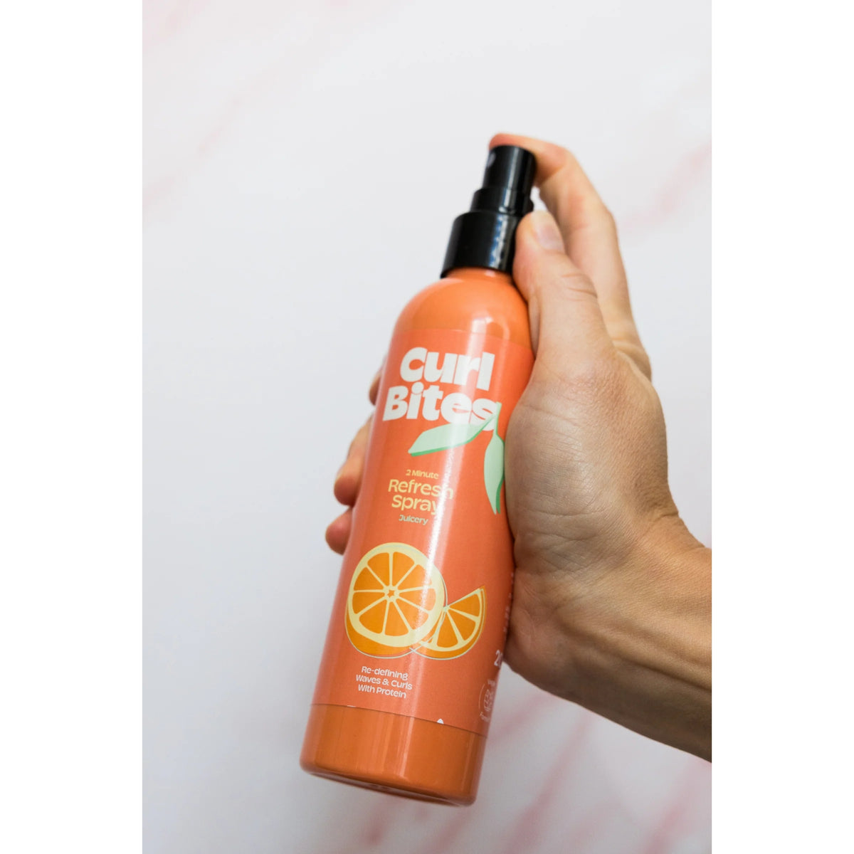 Hånd holder Curl Bites 2 Minute Refresh Spray, orange flaske med citrusfrugter, mod lys baggrund.
