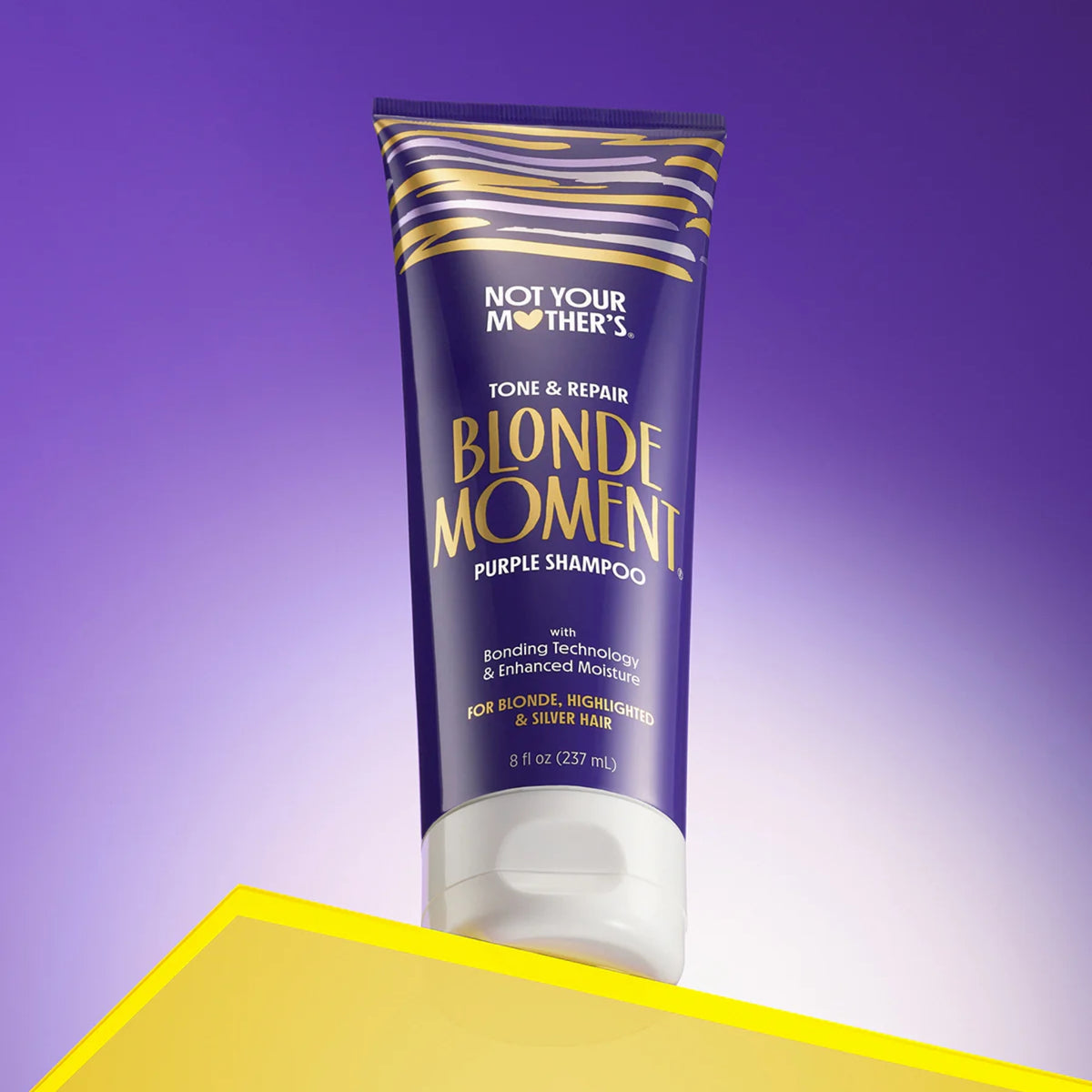 Not Your Mother's Blonde Moment lilla shampoo til blondt hår i lilla og guld tube på gult bord.