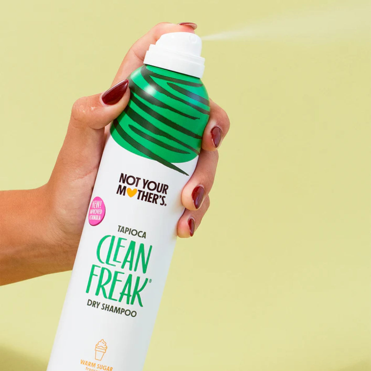 Hånd holder Not Your Mother's Clean Freak tørshampoo i grøn dåse, der sprøjtes mod grøn baggrund.