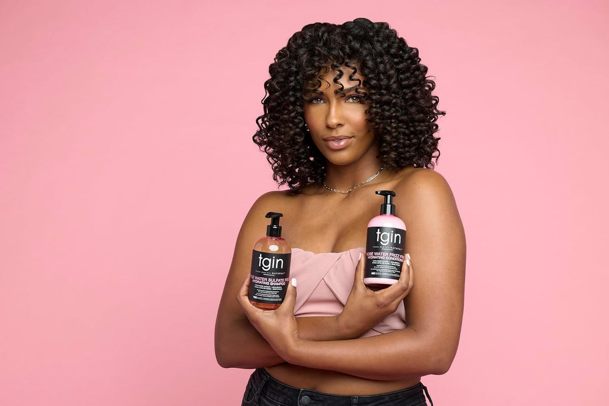 Kvinde med krøllet hår holder TGIN Rose Water Hydrating Leave in Conditioner 384ml foran lyserød baggrund.
